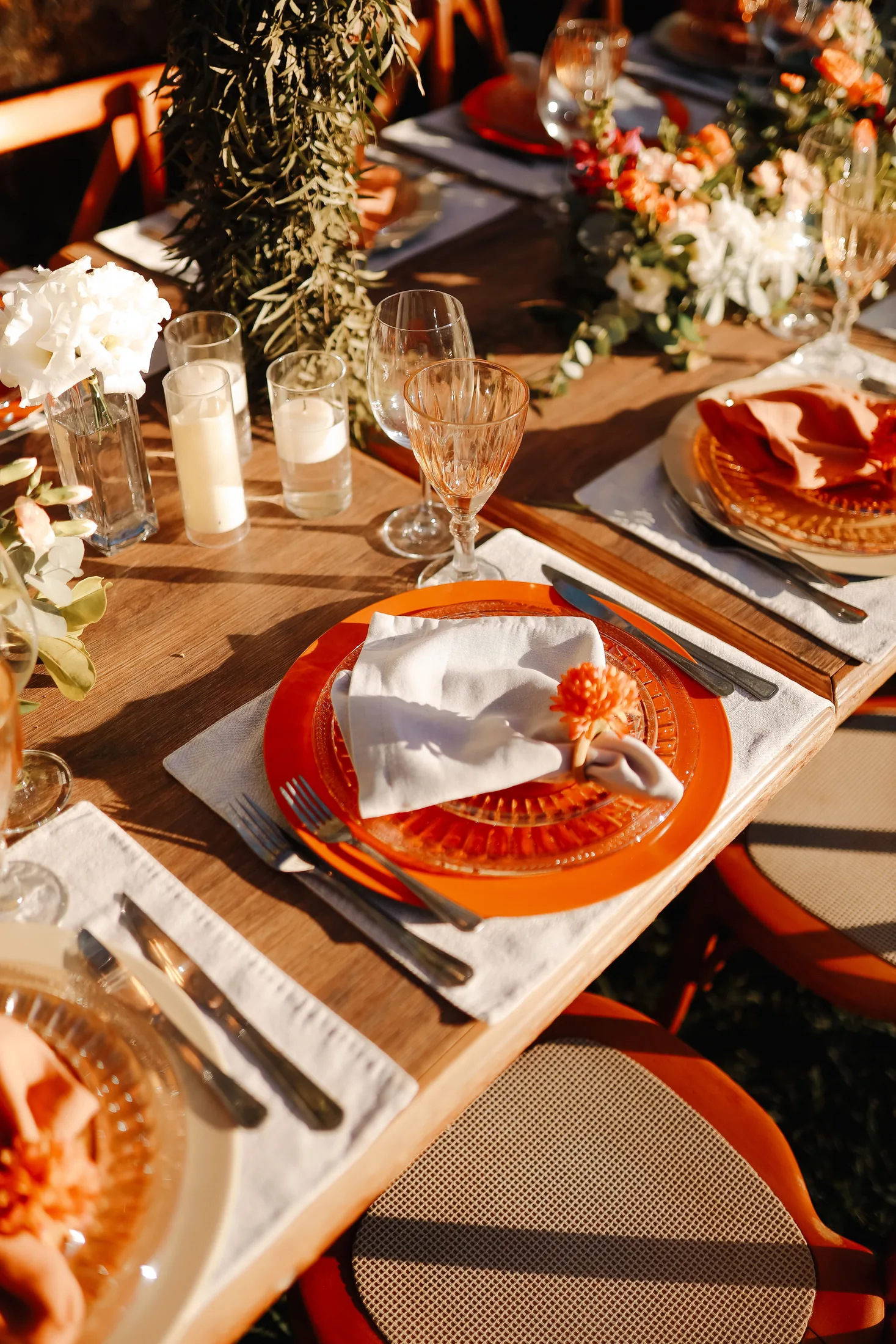 A sunlit dinner table set with orange glassware, flowers and candles